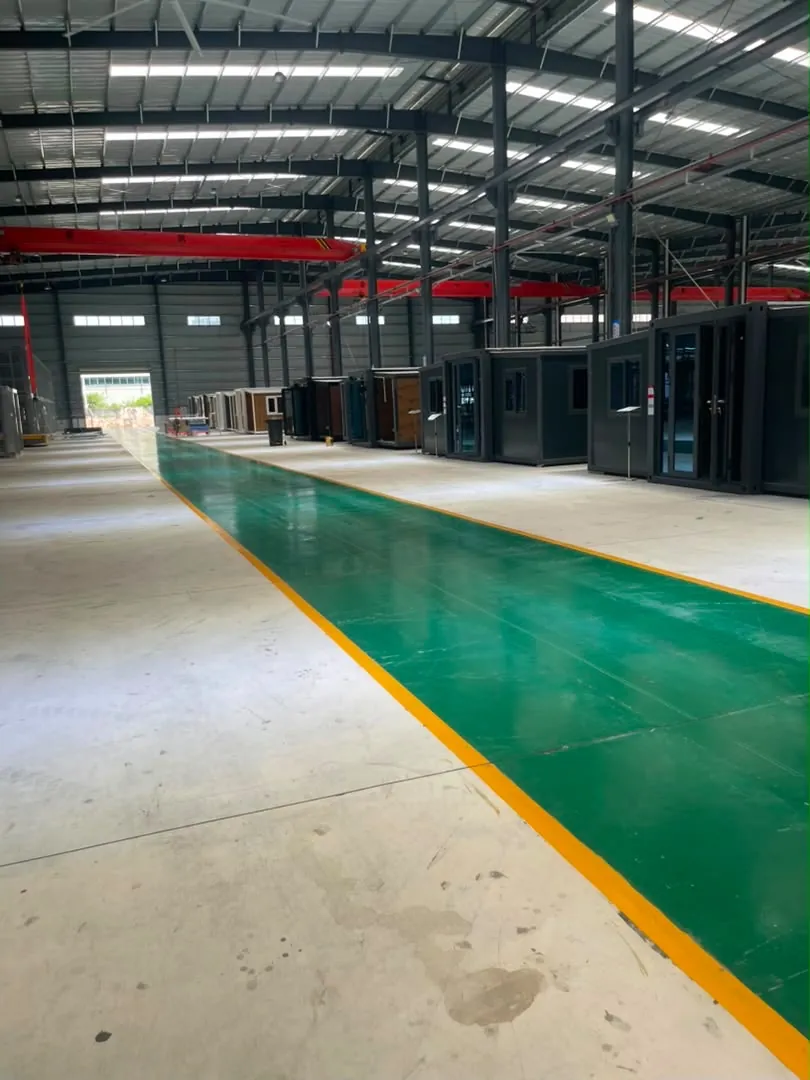 A long green walkway between rows of finished expandable houses under a red overhead crane in the factory hall