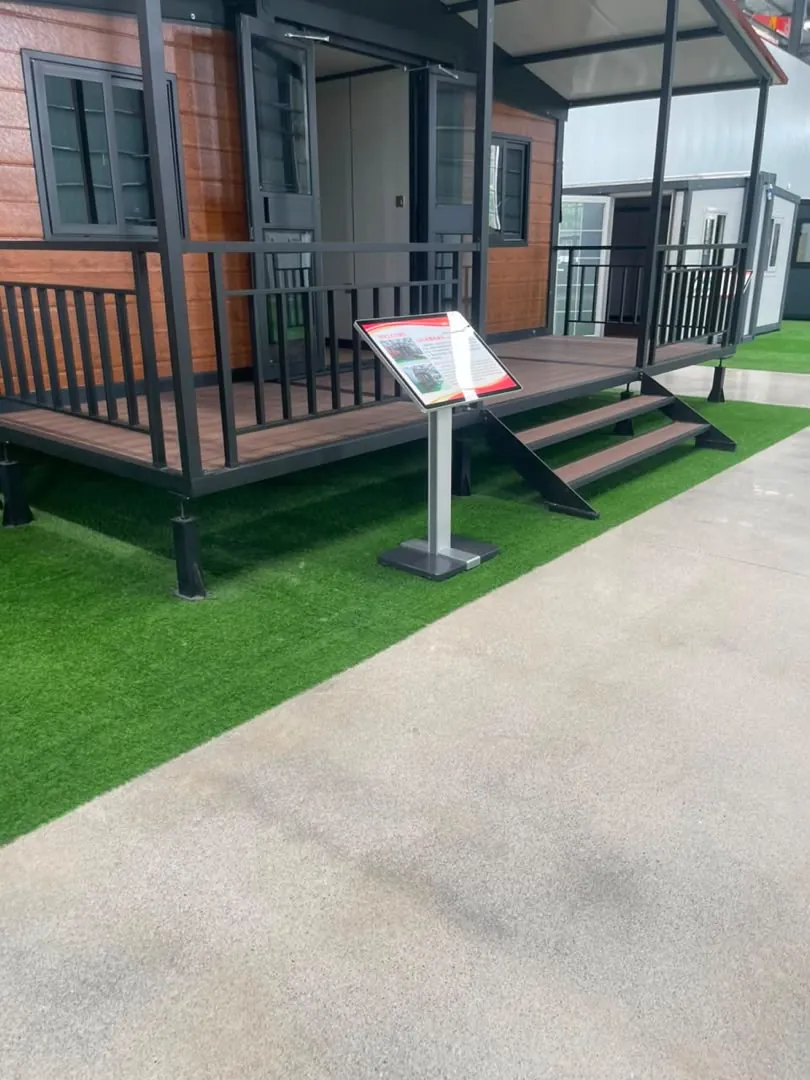 A showroom expandable house at the factory with a roof canopy on black posts, a terrace with railing and entry steps, on artificial grass