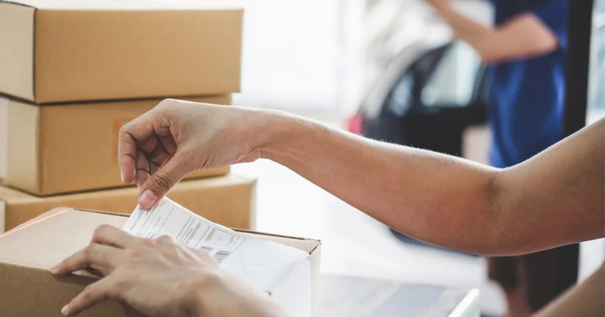 Hands attaching a shipping label to a cardboard parcel with a barcode scanner beside it